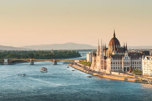 Aerial view of a river with boats, a bridge, and a large historic building with a dome, set against a backdrop of hills and a clear sky.
