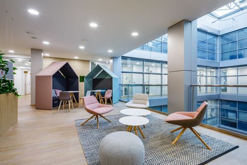 Modern office lounge with pastel chairs, round tables, and geometric seating nooks. Large windows and skylights illuminate the space.