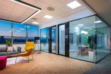 Modern office space with glass-walled meeting rooms, colorful furniture, wooden flooring, and large windows overlooking a cityscape at dusk.