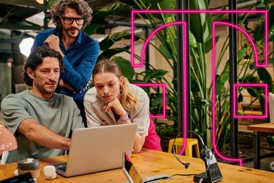 Three people collaborate at a table with a laptop, surrounded by plants and a large magenta illuminated Telekom logo in the background.