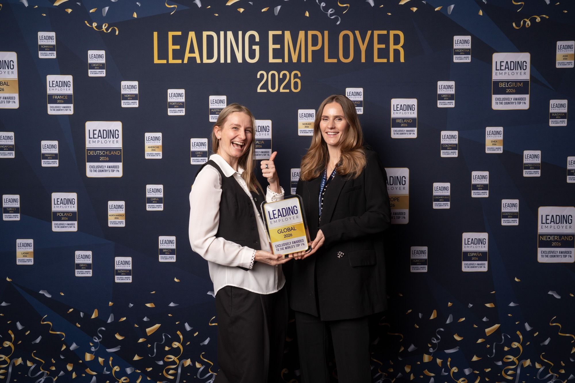 Two women smiling and holding an award in front of a "Leading Employer 2026" backdrop covered with confetti patterns and logos.