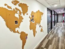 Corkboard world map on office wall with photos pinned on various continents in a hallway with wooden flooring and glass-paneled rooms.