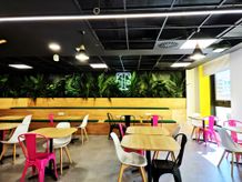 Modern cafe lounge with wooden tables, colorful chairs, potted plants on a bench, and a neon sign on a green wall, under a grid ceiling.