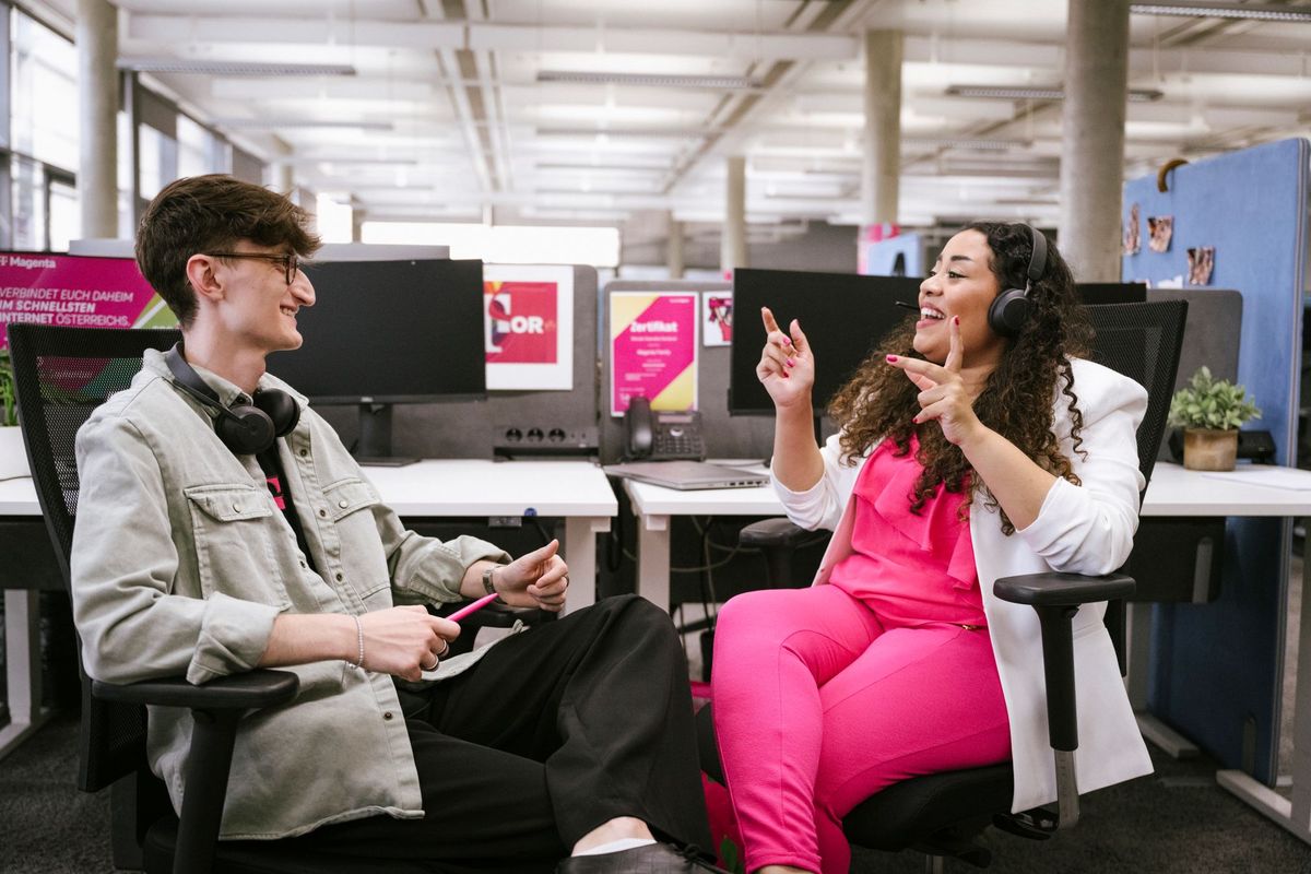 Two colleagues wearing headsets chat and smile in a modern office with computers and colorful decor in the background.