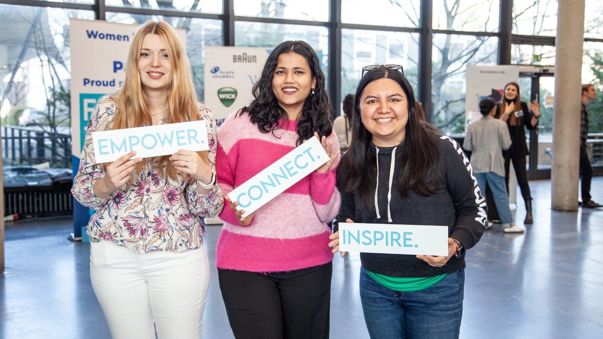 Three women smiling and holding signs reading "EMPOWER," "CONNECT," and "INSPIRE" at an indoor event, with people and banners in the background.