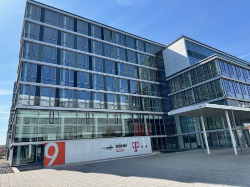 Modern glass office building with multiple floors, featuring company logos on the facade and a clear blue sky in the background.