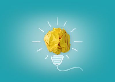 Crumpled yellow paper forms a light bulb shape with drawn lines on a blue background, symbolizing creativity and ideas.