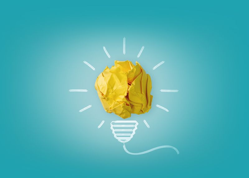 Crumpled yellow paper forms a light bulb shape with drawn lines on a blue background, symbolizing creativity and ideas.