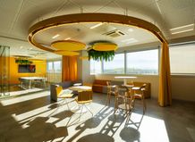 Modern office lounge with circular lighting, yellow accents, round tables, chairs, and large windows letting in natural light.
