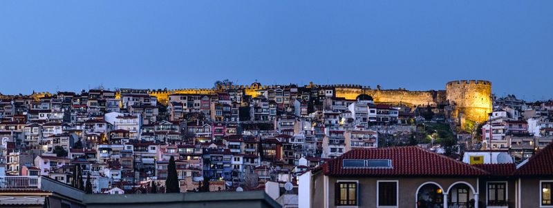 Panoramic view of a hillside cityscape at dusk, featuring illuminated ancient walls and a tower, with densely packed buildings below.