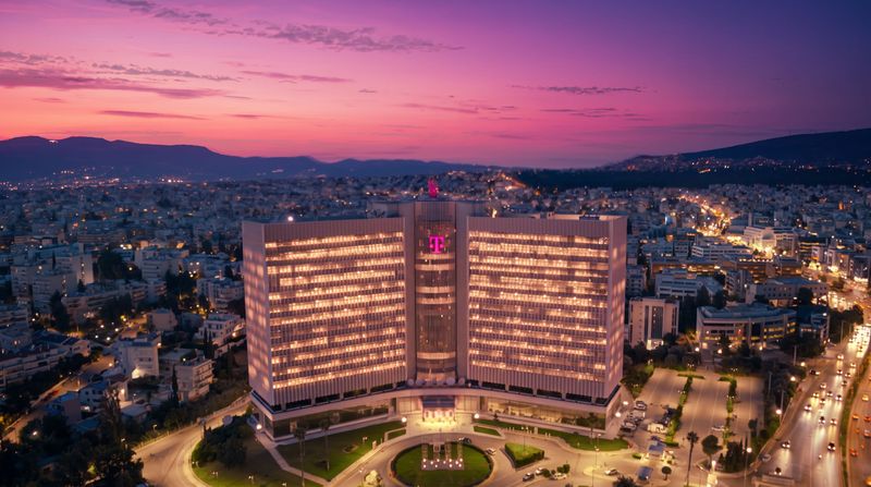 Illuminated cityscape at dusk with a prominent, lit building displaying the magenta T logo, against a vibrant purple and pink sunset sky.