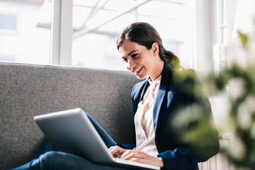 Smiling woman in a blazer sits on a couch using a laptop, with natural light from large windows in the background.