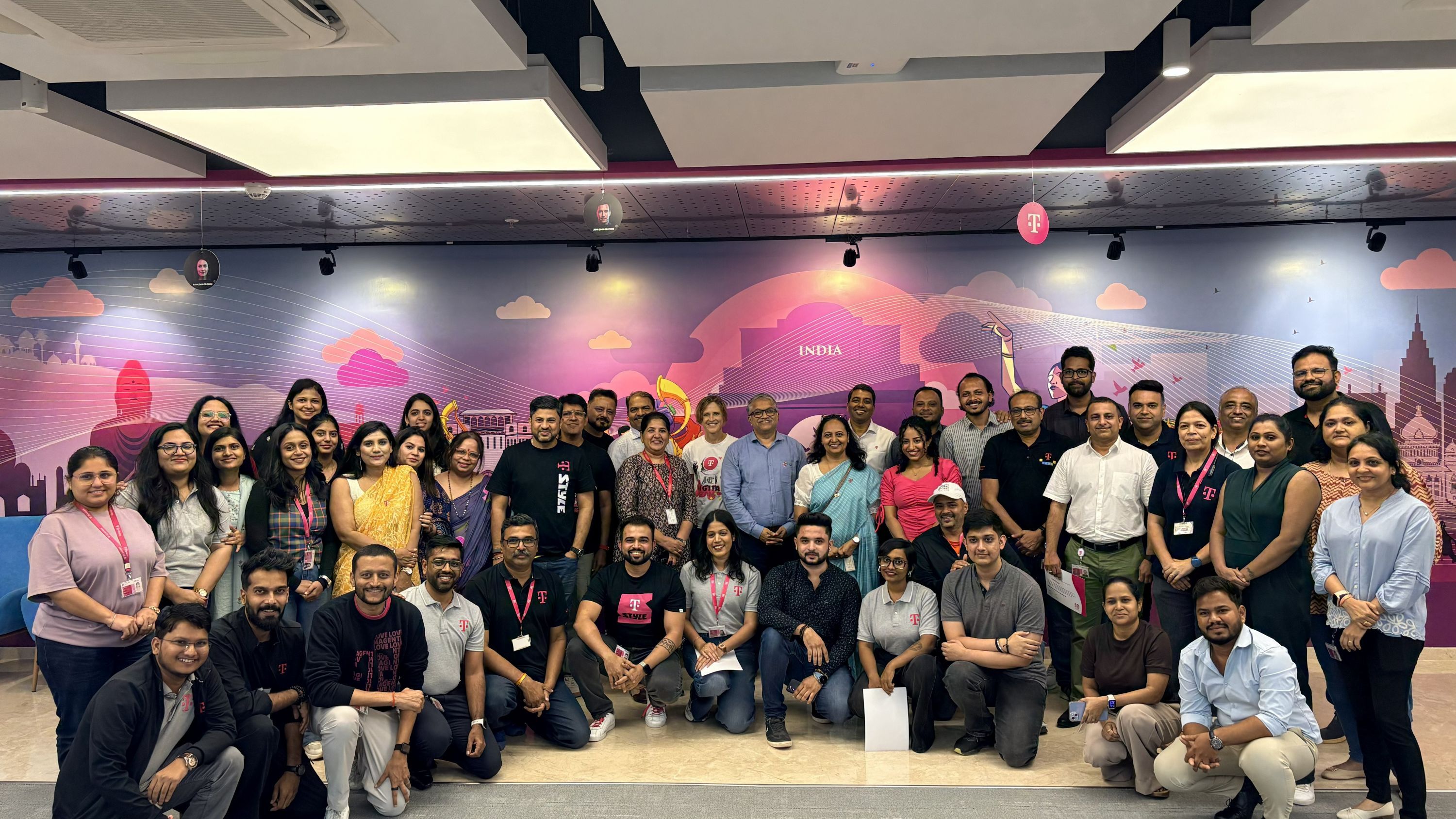 A large group of people smiling and posing together indoors, in front of a colorful mural with an "India" theme.