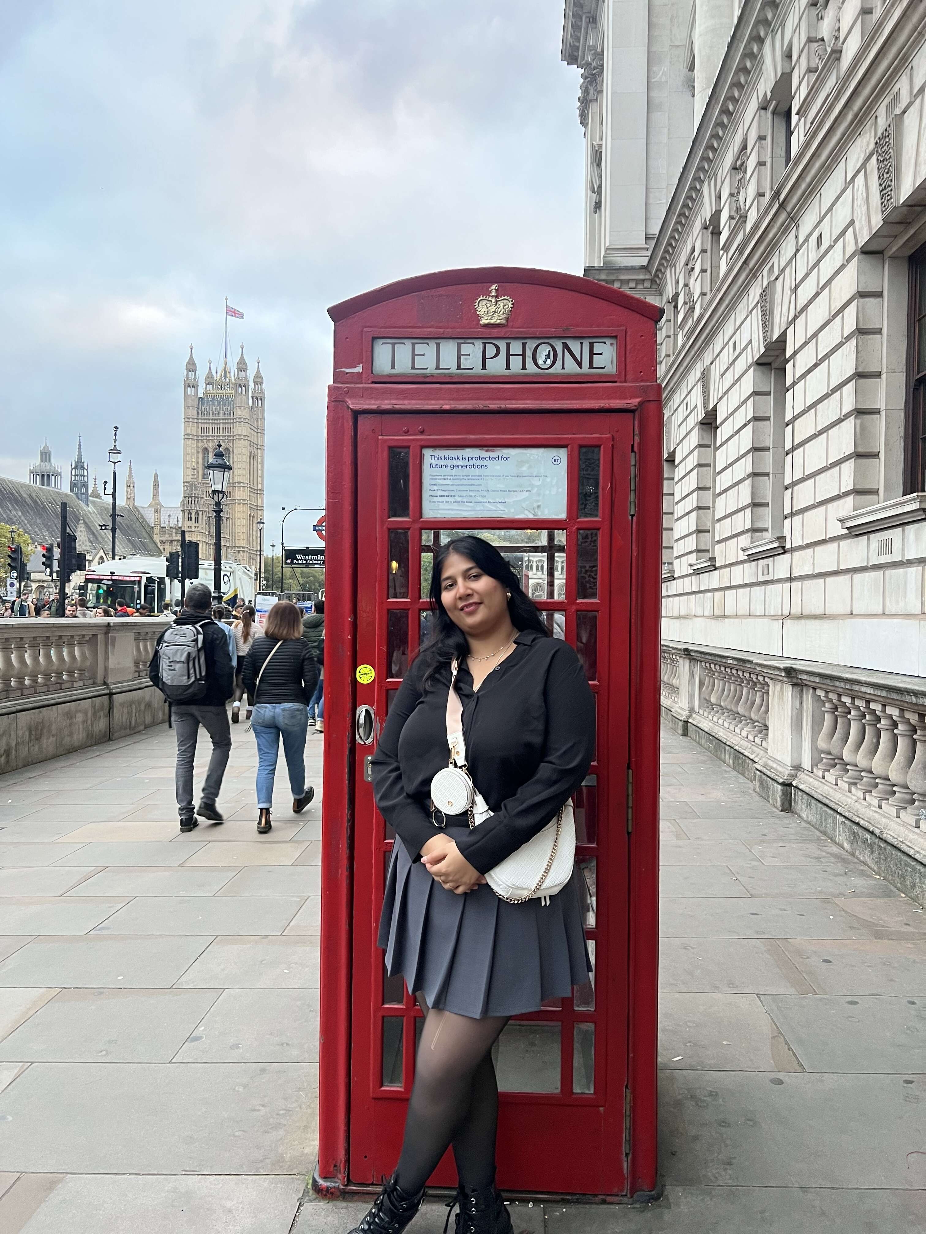 Woman in black top and gray skirt leans against a red telephone booth. Background shows people walking and historic buildings.