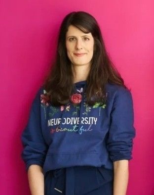 Woman with long hair in a blue sweatshirt with floral embroidery and the phrase "Neurodiversity is Beautiful" stands against a pink background.
