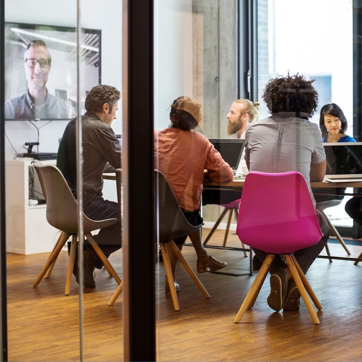 A diverse group of people in a conference room having a meeting, with one person on a video call displayed on a screen.