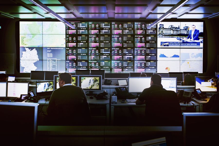 A control room with multiple monitors displaying maps, data, and news. People work at desks with computers, facing large screens on the wall.
