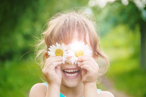 Child smiling with daisies covering their eyes, standing outdoors with a blurred green background.