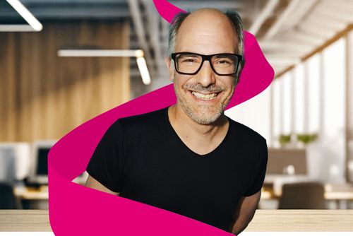 Smiling man with glasses and a black shirt, surrounded by a magenta ribbon, in a modern office setting.