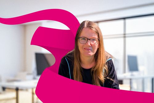 Woman with glasses and long hair smiles in a bright office. A magenta ribbon overlaps the image.