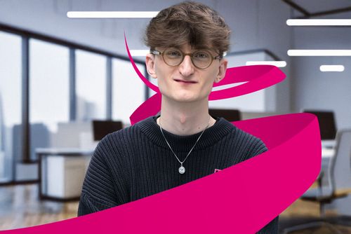 Person with glasses and curly hair smiling, wearing a black sweater and necklace. Magenta ribbon design overlays the image. Office setting in the background.