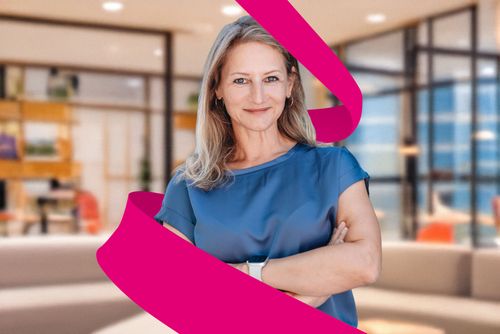 Woman with crossed arms, wearing a blue blouse, stands confidently in a modern office with a magenta ribbon graphic in the foreground.