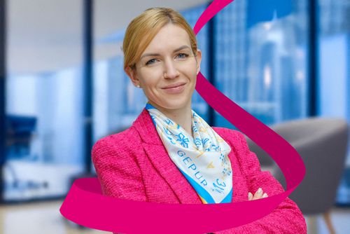 Smiling woman in a magenta blazer and patterned scarf, standing confidently in a modern office with large windows and city view.