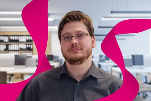 A person with glasses and a beard in a gray shirt against an office background, with magenta abstract shapes in the foreground.
