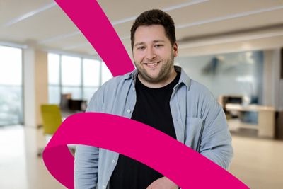 Smiling man wearing a blue shirt and black t-shirt stands in a modern office with a magenta ribbon effect in the foreground.