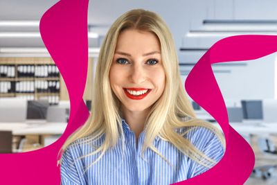 Smiling woman with long blonde hair in a blue striped shirt, standing in a modern office with pink abstract shapes in the foreground.