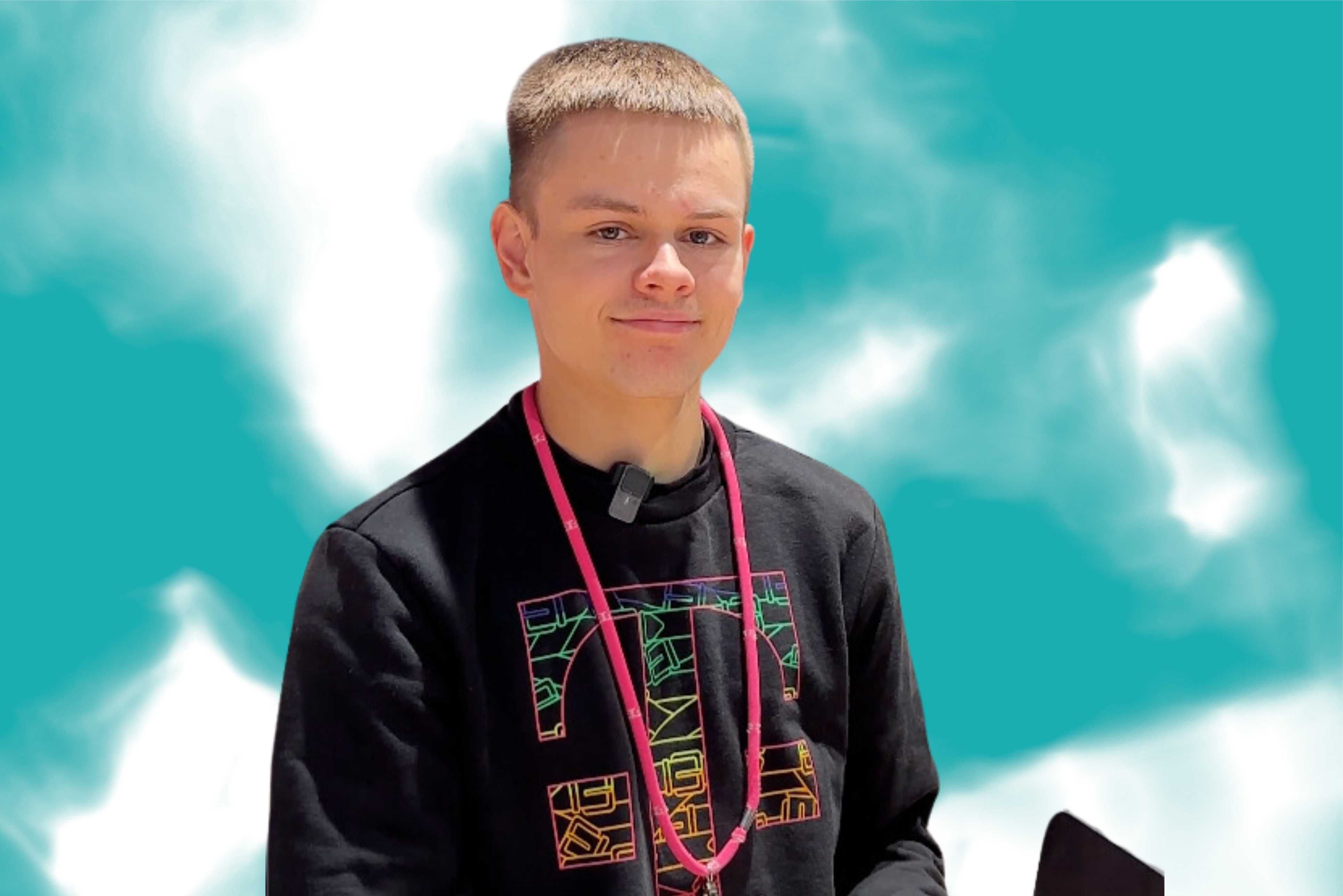 Young boy with short hair, wearing a black shirt with colorful geometric designs and a pink lanyard, against a cloudy blue sky background.