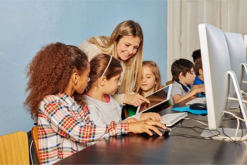 A teacher helps a group of children using computers and tablets.