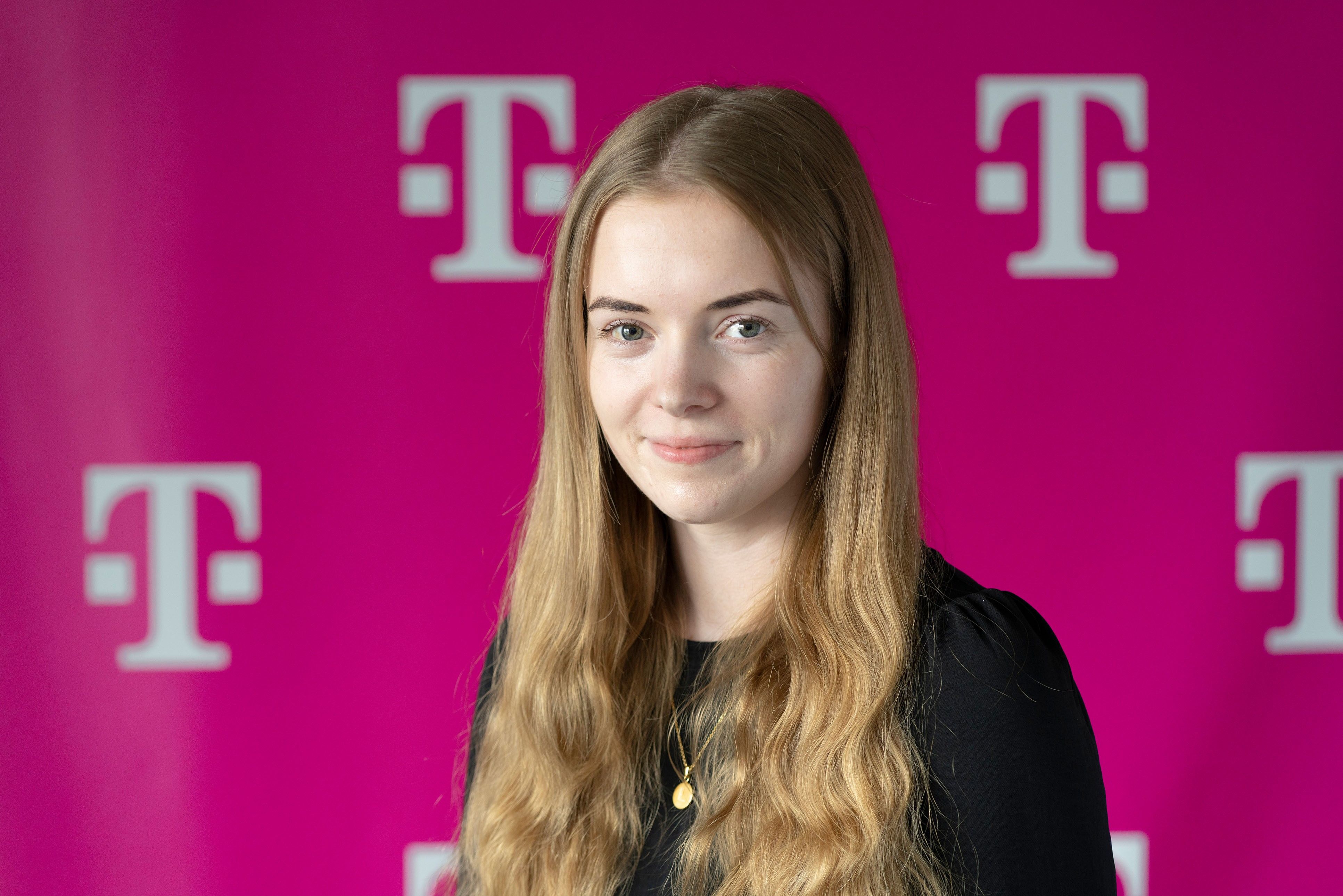 A person with long blonde hair, wearing a black top, smiles against a pink background with white "T" logos.