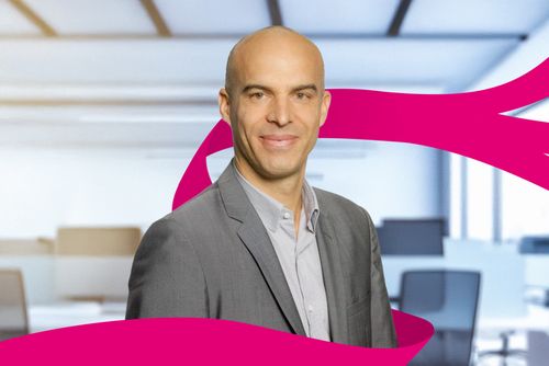 Bald man in a gray suit smiling, standing in a bright office with a pink ribbon-like design in the foreground.