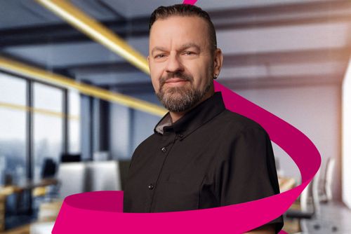 Man with a beard and short hair in a black shirt, standing in a modern office with a pink ribbon swirling around him.