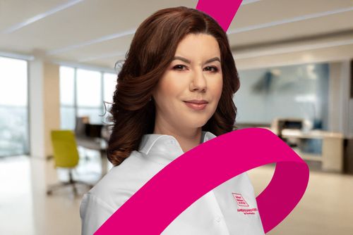 Woman with brown hair in a white shirt stands in a modern office, with a pink ribbon graphic in the foreground.