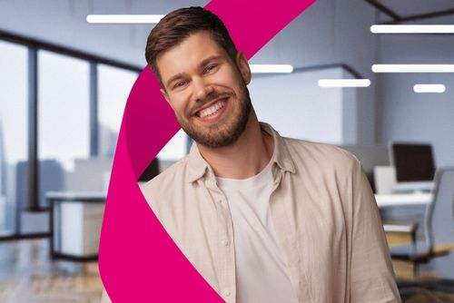 Smiling man with a beard in a light shirt stands in a modern office with large windows and a pink abstract shape in the foreground.