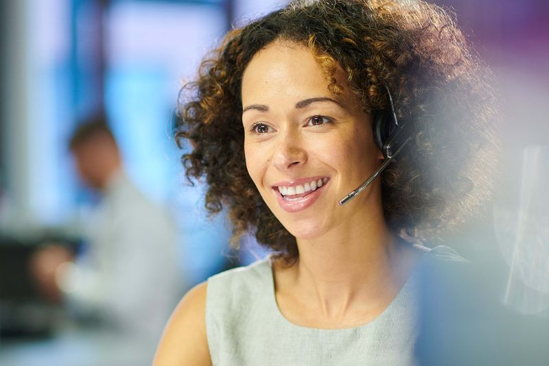 Smiling woman with curly hair wearing a headset, sitting in an office environment, appears to be engaged in a conversation.