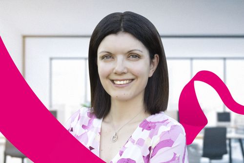 Smiling woman with dark hair in a pink and white blouse, standing in a modern office setting with a pink ribbon design overlay.