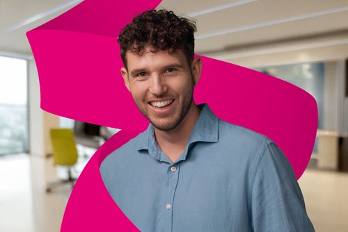 Smiling person with curly hair in a blue shirt, standing in a modern office with a bright magenta abstract shape in the background.