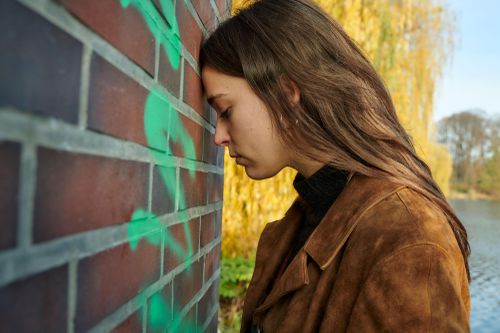 Woman in a brown jacket leans her head against a graffiti-covered brick wall, outdoors, with autumn trees and a river in the background.