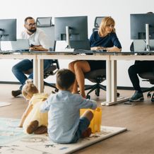 Children playing on the floor while adults work at computers in an office setting.