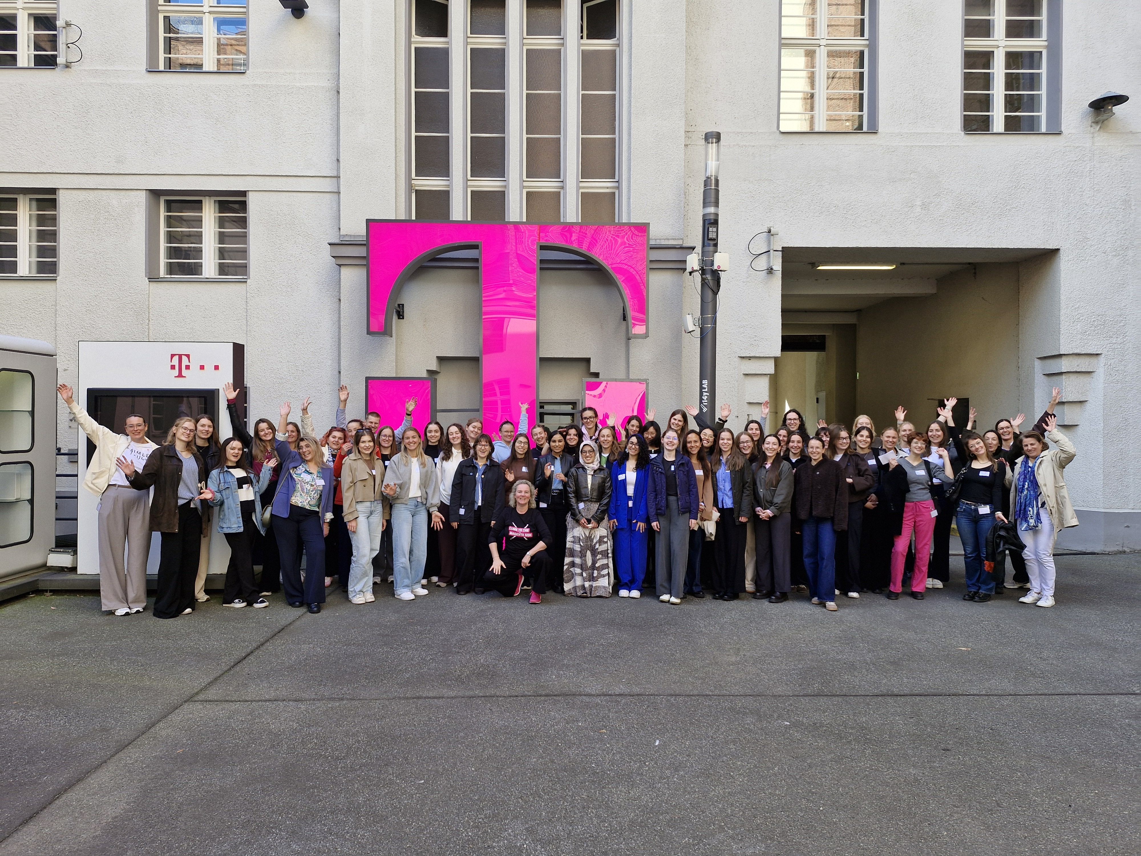 A group of people smiling and waving in front of a large pink logo on a building.