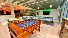 Modern recreation room with foosball and ping pong tables, colorful seating, potted plants, and vibrant ceiling lights.