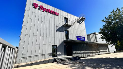 Modern gray building with "T-Systems" and "REDESSA INNOVACIÓ" signs, fire escape stairs, and a clear blue sky in the background.