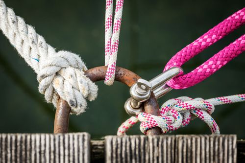 Close-up of various ropes, including white and pink, tied to metal rings and secured with a shackle on a wooden surface.