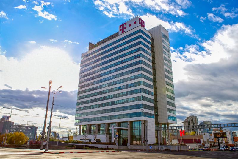 Modern multi-story office building with glass windows, under a partly cloudy sky, surrounded by urban elements and roads.