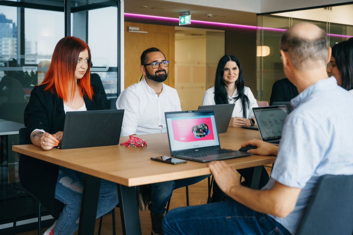 Five people in a meeting room sitting around a table with laptops, engaged in a discussion.
