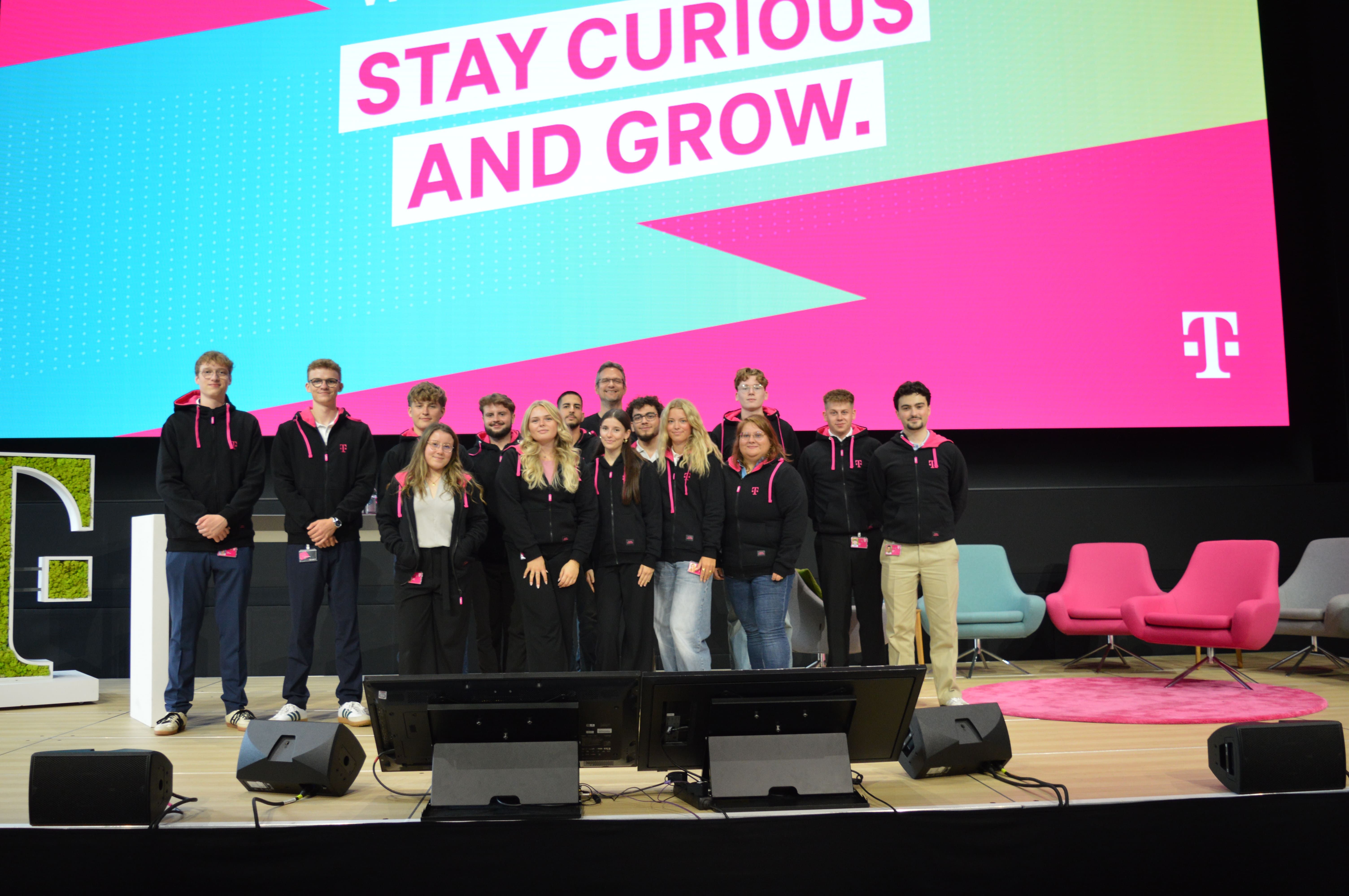 A group of people in matching attire pose on stage in front of a screen displaying "Stay Curious and Grow."