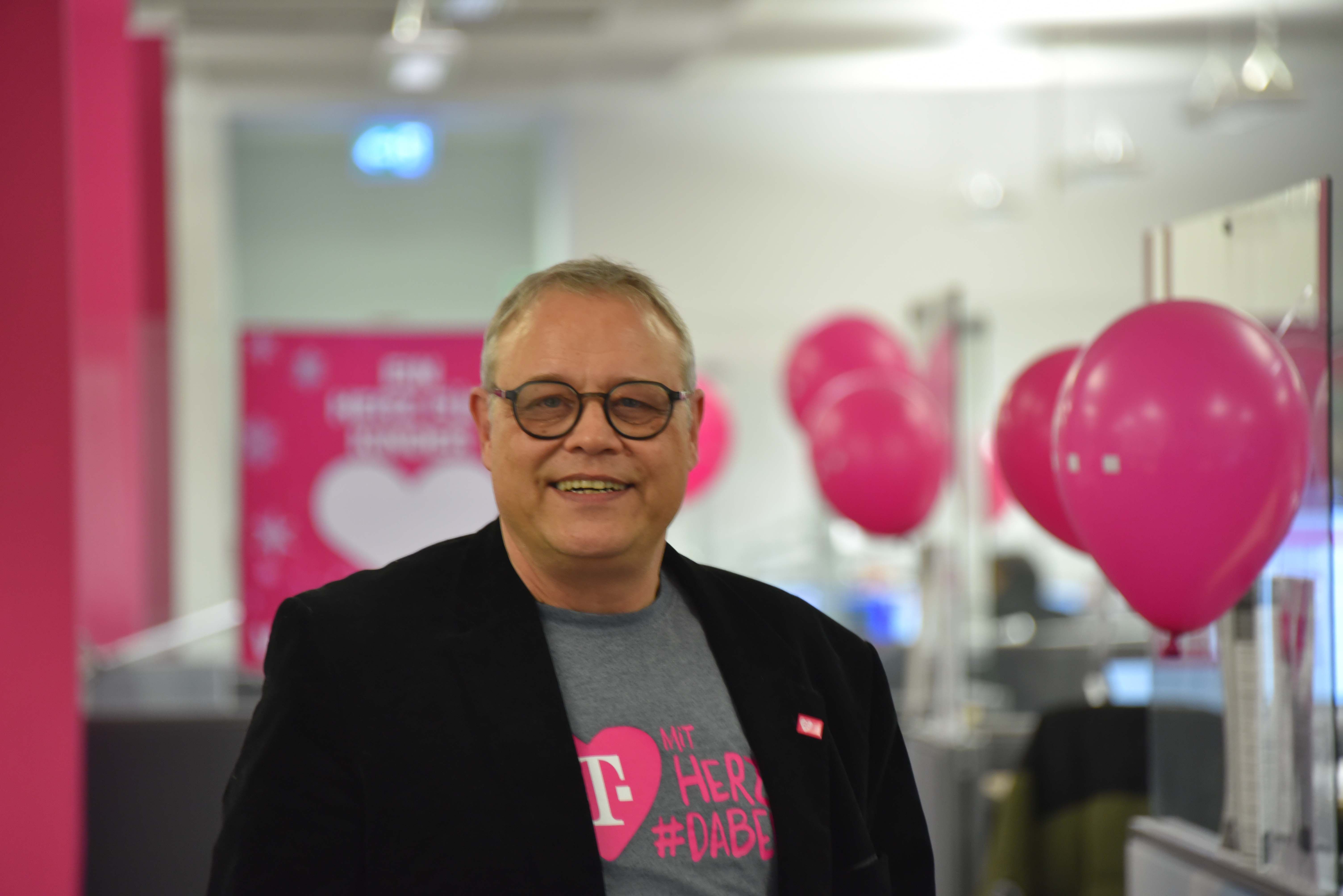A smiling man wearing glasses and a dark blazer stands in an office with pink balloons and a pink-themed poster in the background.